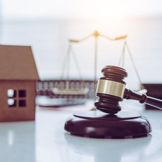 Auf einer Tischplatte steht ein Modellhaus, eine Waage, die Gerechtigkeit symbolisiert und der Hammer eines Richters, um ein Urteil zu fällen. Oft führen Testamente zu Erbstreitigkeiten, zum Beispiel, wenn Kinder enterbt oder Stiefkinder nicht bedacht werden. Nicht alle haben einen Anspruch auf einen Pflichtteil des elterlichen Vermögens.
