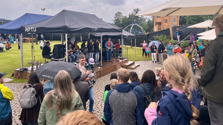 Kinder singen auf einer Bühne vor einem Publikum, das im Regen steht: Der Unisport der Rheinland-Pfälzischen Technischen Universität Kaiserslautern-Landau (RPTU) hat auf dem Campus einen Abenteuerspielplatz eröffnet. Das Kitz-Land können Kinder mit einer Unisport-Karte besuchen.