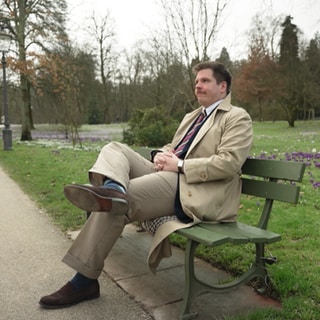 Timo Wolfinger sitzt auf einer Parkbank. Er trägt einen Trenchcoat. 