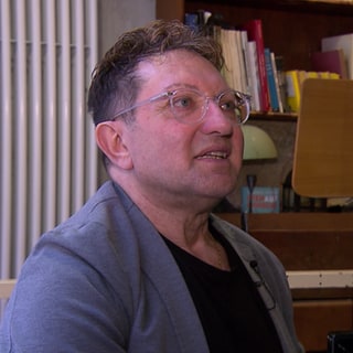 Pförtner Enzo trägt eine Brille und sitzt an einem Klavier.