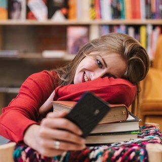 Studentin liegt mit dem Oberkörper auf einem Stapel Bücher und schaut froh in ihr Smartphone: Studierende scheitern oft an langen Texten. Klagen über die Leseschwäche an den Universitäten ziehen sich quer durch die Fächer. Ist das wirklich so schlimm? 