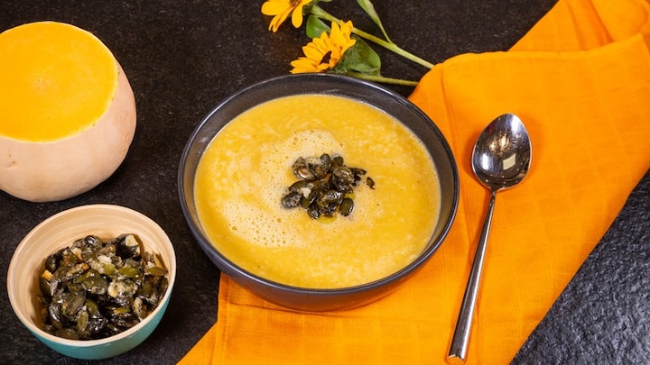 Die fertige Kürbissuppe auf einem schwarzen Tisch mit orangener Tischdecke. Sonnenblumen, ein aufgeschnittener Butternut-Kürbis und die karamelisierten Kürbiskernen stehen als Deko neben dem Teller.