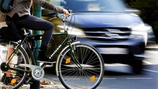 Ein Radfahrer wartet an einer Ampel, ein Auto fährt vorbei (Symbolbild). In RLP kommt es im Straßenverkehr offenbar immer öfter zum Streit zwischen Autofahrern und Radfahrern.
