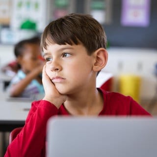 Ein Junge sitzt traurig in seiner Schulklasse und schaut abwesend in die Ferne. Zu oft werden Signale der Gewalt an Kindern nicht erkannt – auch nicht von jenen, die in Kitas, Schulen, Arztpraxen und Jugendämtern mit ihnen zu tun haben.