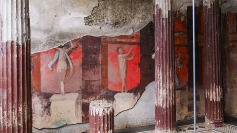Fresko des Dionysos-Kults in Pompeji mit rötlichen Wandmalereien zwischen antiken Säulen, das rituelle Szenen der dionysischen Mysterien zeigt.