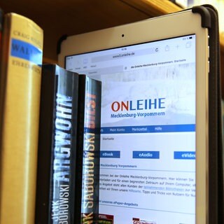 Ein iPad mit der Internetseite der Onleihe Mecklenburg-Vorpommern steckt zwischen den Büchern im Regal einer Bibliothek: Autoren und Verlage sehen ihre Existenz durch die sogenannte Onleihe gefährdet, Bibliotheken beklagen, dass ihnen Bestseller oft erst Monate nach Erscheinen verkauft werden. 