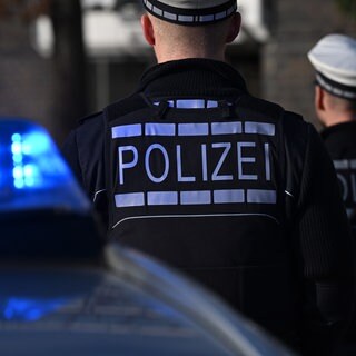 Das Logo der Polizei auf der Dienstkleidung von Polizisten die vor einem Blaulicht stehen (gestellte Szene).