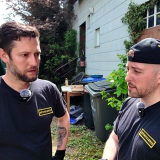 Zwei Männer, die als Entrümpler arbeiten, stehen vor einem alten Haus, neben dem ein Container steht.