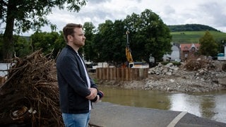 Marius Reichert trägt ein weißes T-Shirt und ein dunkles Blouson, er hält ein Mikrofon in den Händen. Er steht an einem ruhigen Fluss und schaut mit nachdenklichem Gesichtsausdruck nach rechts. Im Hintergrund sieht man eine von der Flutwelle zerstörte Brücke, viel Schutt und Äste an beiden Seiten des Flussufers.