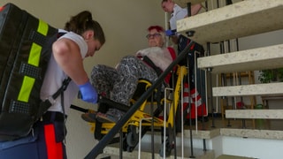 Die zwei Notfallsanitäterinnen Alina Peschers und Stephanie Dahlem tragen eine ältere Dame in einem Rettungsstuhl eine Treppe runter.