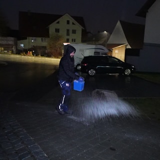 Auszubildender Amir ist mit einer schwarzen Jacke bekleidet. Er läuft nachts über eine Straße und streut Taumittel aus einem Eimer auf den Boden.