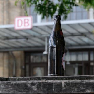 Eine Bierflasche steht vor einem Bahnhof auf einer Bank. Die Deutsche Bahn plant einen radikalen Schritt mit der Hoffnung auf mehr Sicherheit für die Fahrgäste und die Beschäftigten.