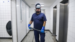 Sinan steht mit einem Hockdruckreiniger in der weiß gefliesten Toilette am Charlottenplatz. Er hat eine große, kräftige Statur, und trägt ein blaues Shirt und einen Helm mit Gesichtsschutz. 