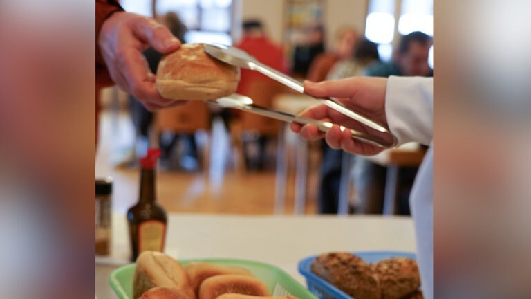 Ein Brötchen wird mit einer Zange einer Hand gereicht.