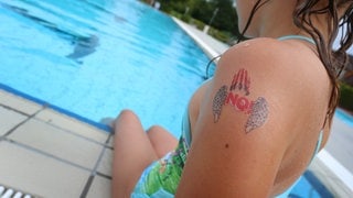 Ein Mädchen mit einem "NO!" Klebetattoo am Arm sitzt im Freibad am Beckenrand. 