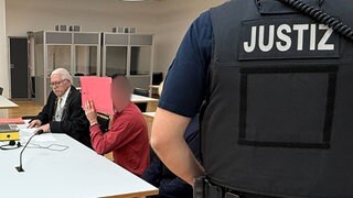 Im Gerichtssaal sitz der Angeklagte neben seinem Anwalt. Er hält sich eine rötliche Mappe vor das Gesicht. Am Rand steht ein Justizbeamter.