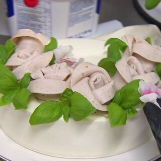 Eine weiße Torte mit weißen Wurströschen, grüner Basilikum-Garnitur und einer Frischkäseglasur. Von rechts kommt ein schwarzer Handschuh, der eine Blüte draufsetzt.