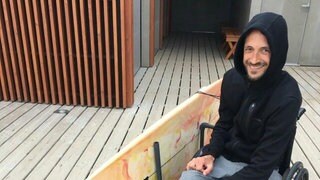 Der querschnittgelähmte Johannes Laing sitzt lächelnd in seinem Rollstuhl, an dem mit Hilfe einer selbstgebauten Halterung sein Surfbrett befestigt ist.