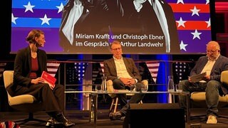 Drei Menschen sitzen vor einem überdimensionalem Bild. Auf diesem sind die beiden Kandidaten für die kommende US-Wahl zu sehen. Links Kamala Harris und rechts Donald Trump.