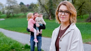 Jenny steht auf einem asphaltierten Weg und schaut in die Kamera. Weiter hinten steht ihr Ehemann Sascha, der beide Töchter in den Armen hält. Im Hintergrund sind Bäume und eine Wiese zu sehen.