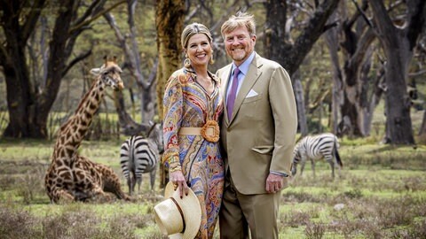 König Willem-Alexander und Königin Máxima stehen in der kenianischen Natur vor Giraffen und Zebras und lachen in die Kamera.