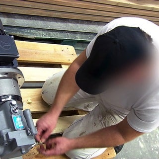 Insasse der JVA Mannheim arbeitet im Malerbetrieb. Er kniet auf einer Palette und arbeitet an einer Maschine.