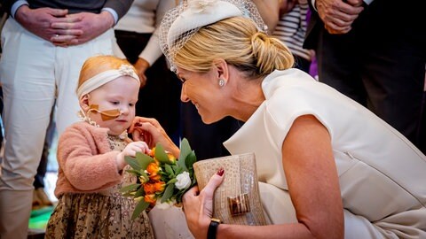 Königin Máxima in weißem Outfit mit einem krebskranken Mädchen, das ihr einen Blumenstrauß übergeben hat