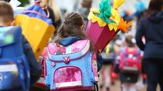 Zwei Kinder, die von hinten fotografiert sind, sind mit Schulranzen und Schultüte auf dem Weg zur Einschulung.