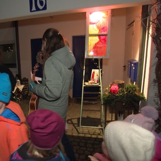 Vor einem Haus stehen viele Kinder und einige Erwachsene. Ein erleuchtetes Fenster hängt über einer Leiter. Das Fenster ist bunt bemalt.