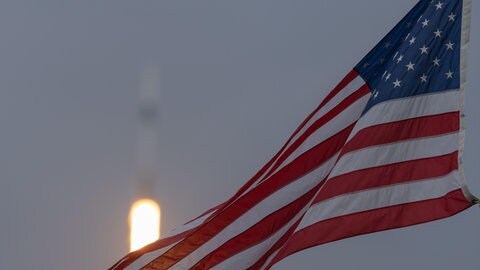 Amerikanische Flagge weht, im Hintergrund startet eine Rakete. Die Europäische Weltraumorganisation ESA plant den Ausbau ihrer unabhängigen Raumfahrtkapazitäten.