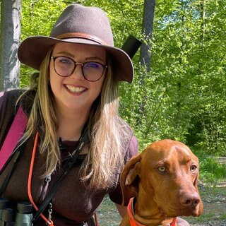 Isabel Berger sitzt in einem Wald auf dem Boden und legt ihren linken Arm um ihren braunen Jagdhund Eros.