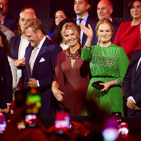 Eine lächelnde Frau in braunem Kleid, Königin Màxima, steht während einer Feierlichkeit neben einer jungen Frau in grünem Kleid, die winkt, Tochter Amalia.