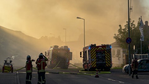 Feuerwehrleute und Feuerwehrautos stehen in Karlsruhe im Stadtteil Mühlburg. Rauchwolken steigen in den Himmel.