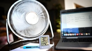 Ein Thermometer an einem Arbeitsplatz zeigt eine Temperatur von über 30 Grad Celsius. Daneben steht ein Ventilator.