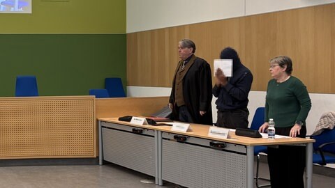 Der Angeklagte (Mitte) zu Beginn des letzten Verhandlungstags im Prozess versuchter Mord. Auch bei der Urteilsverkündung, als es um Femizid ging, hielt er sich wieder einen Zettel vor das Gesicht.