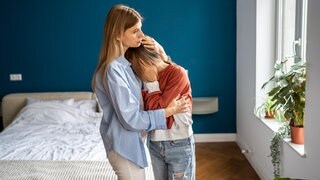 Mutter umarmt tröstend die weinende Tochter zu Hause | Früher Tod eines Elternteils: Wie lassen sich Kinder in ihrer Trauer begleiten?