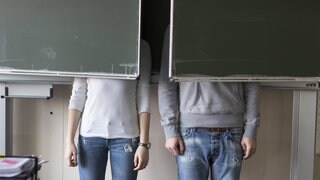 Zwei Schüler stehen hinter einer Tafel im Klassenzimmer, ihr Oberkörper ist von der Tafel verdeckt.