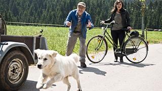 Karl ruft Hofhund Willy hinterher, der wegrennt, Eva steht mit ihrem Fahrrad neben Karl