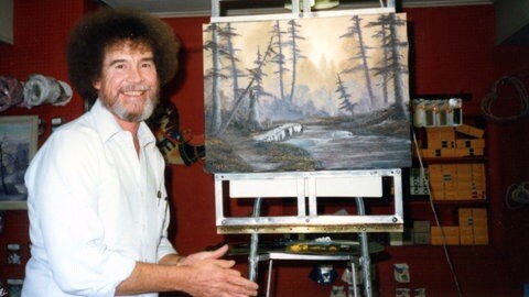 Bob Ross sitzt lächelnd vor einer gemalten Landschaft