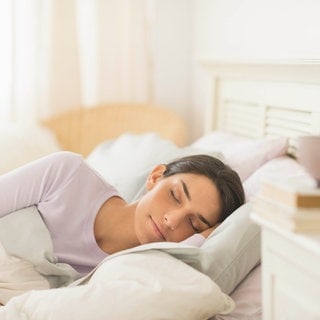 Eine junge Frau liegt seitlich im Bett und schläft entspannt: Dauerhaft schlecht und zu wenig zu schlafen, schadet Gehirn und Gesundheit. Schlaf lässt sich zwar nicht erzwingen, doch er lässt sich erlernen und ist sowieso meist besser, als viele meinen.