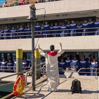 Viele Jugendliche stehen auf einem Schiff an der Reling. Sie tragen einheitliche Gewänder. Im Vordergrund, auf dem Festland, steht auch jemand in Gewand und dirigiert. 