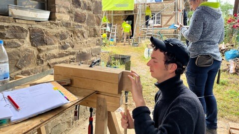 Ein junger Handwerker sägt an einem schmalen Holzbrett. Vor ihm sieht man ein altes Gemäude.