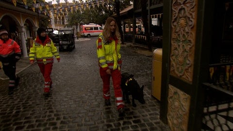Hundeführerin Laura Goebes läuft in geld-roter Rettungsjacke durch den Europa-Park. An der Leine einer der Rettungshunde, die hier heute trainieren: ihre schwarze Schäferhündin Ombra. Im Hintergrund zwei Kollegen in Rettungskleidung.