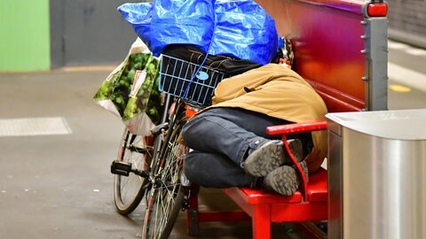 Wohnungslose Person liegt auf einer roten Holzbank und schläft. Die Zahl der wohnungslosen Menschen ist in den letzten Jahren auch in RLP gestiegen. 