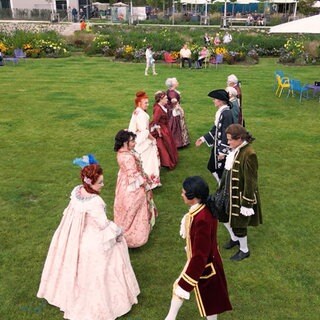 Eine Gruppe von Leuten steht auf einer Wiese. Alle tragen Kleidung im barocken Stil mit bunten Farben.