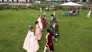 Eine Gruppe von Leuten steht auf einer Wiese. Alle tragen Kleidung im barocken Stil mit bunten Farben.