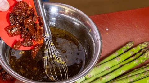 Kleingeschnittene getrocknete Tomaten werden mit einer Hand von einem roten Teigschaber in eine Schüssel mit bräunlicher Flüssigkeit gegeben.
