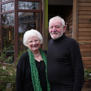 Frau und Mann mit weißen Haaren stehen lächelnd vor Holzhauseingang.