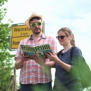 Florian Ladenburger und Laura Bieber stehen mit einem Rom-Reiseführer vor dem Ortsschild von Rommelshausen Richtung Weistadt.