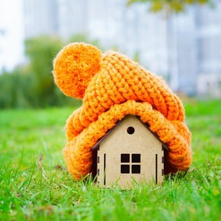 Holzhäuschen mit dicker orangefarbener Strickmütze auf dem Dach (Symbolbild) auf einer Wiese: Viele Wohnungen sind noch immer schlecht isoliert, Besitzer und Mieter heizen wortwörtlich zum Fenster hinaus; die energetische Sanierung kommt kaum voran. 